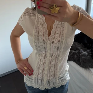Vit blus med spetsdetaljer - Säljer en söt vit blus med v-ringning och vackra spetsdetaljer längs framstycket och ärmslut. Blusen har korta ärmar och en feminin passform som passar perfekt till jeans eller kjol.