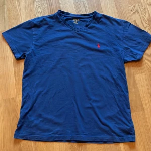 Blå t-shirt från Polo Ralph Lauren - Säljer en snygg blå t-shirt från Polo Ralph Lauren i storlek S med deras röda logga broderad på bröstet. T-shirten har en v-ringad hals och korta ärmar. Perfekt för sommaren! 