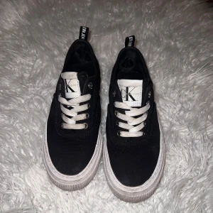 Svarta sneakers från Calvin Klein - Snygga svarta sneakers från Calvin Klein med vit sula och vita snören. Skorna har märkets logga på plösen och sidan. Perfekta för en stilren look.