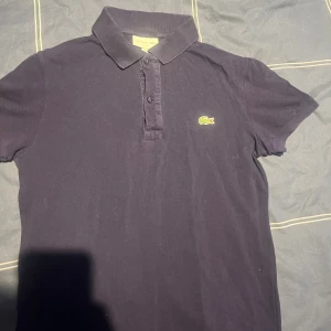 Mörkblå pikétröja från Lacoste - Säljer en klassisk mörkblå pikétröja från Lacoste med den ikoniska krokodilloggan på bröstet. Nått år gammal därav billigt pris, hör av er för fler frågor