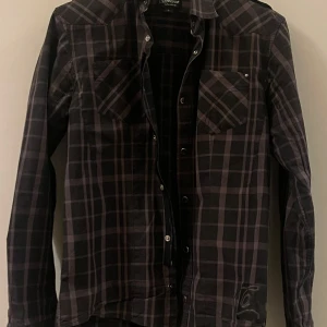 Svart och grå rutig skjorta från Osiris - Snygg svart och grå rutig skjorta från Osiris med långa ärmar och knappar framtill. Skjortan har två bröstfickor med lock och en klassisk krage. Perfekt för en stilren look.