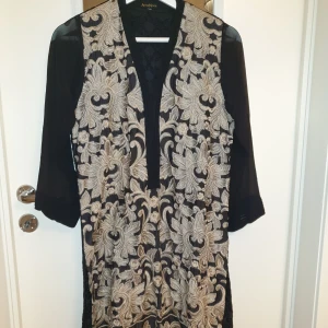 Svart och guld broderad klänning från Agha Noor - Elegant svart klänning med detaljerat guld broderi framtill från Agha Noor. Klänningen har trekvartsärmar och en v-ringad design, slit på sidorna. Perfekt för en stilfull look med sina intrikata mönster och lyxiga känsla.