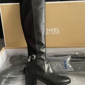 Michael Kors - svarta stövlar   - Snygga svarta stövlar från Michael Kors med dragkedja och klack. Perfekta för en stilren look. Stövlarna har en elegant design med en dekorativ spänne på sidan. Stövlar ovan knä. Material: Läder & Elastisk foder. En mångsidig stil som enkelt kompletterar en hel del bilder. Dessa knähöga stövlar är gjorda av fullnarvsläder och har en elastisk baksida panel för flexibel passform. För ytterligare skydd har hälkappor lagts till.