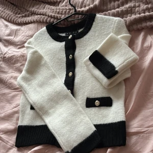 Vit och svart kofta från Amisu - Säljer en stilren vit och svart kofta från Amisu. Koftan har svarta detaljer vid knappar och fickor samt ribbade muddar. Perfekt för en elegant look.