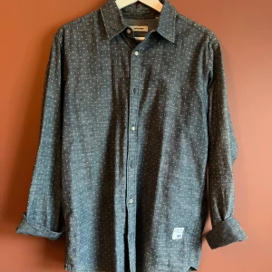 Mörkblå prickig skjorta från Jack & Jones - Snygg mörkblå skjorta från Jack & Jones med små vita prickar. Skjortan har långa ärmar och knappar framtill. Storlek L