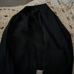 Svart tröja från Massimo Dutti - Snygg svart jacka från Massimo Dutti med en stilren design. Jackan har en dragkedja framtill och är perfekt för en elegant look. Den är tillverkad i ett mjukt material som ger en bekväm passform.