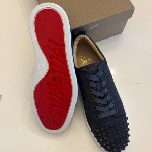 Mörk gråa, Christian Louboutin skor!! - Snygga svarta sneakers från Christian Louboutin med ikoniska röda sulor. Skorna har en unik design med nitar på tån och är tillverkade i skinn. Perfekta för att ge din stil en edgy touch.
