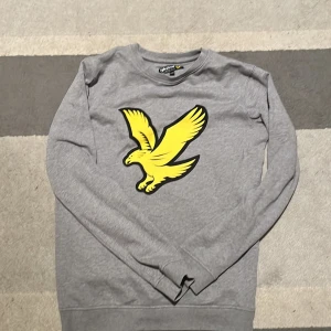 Grå tröja med gul fågel från Lyle & Scott - Säljer en grå tröja från Lyle & Scott med en stor gul fågel på framsidan. Tröjan har långa ärmar och är perfekt för en avslappnad stil. Passar bra till både jeans och joggers.