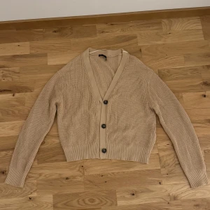Beige kofta med knappar - Säljer en stilren beige kofta med knappar framtill. Den har en ribbad struktur och långa ärmar, perfekt för lager-på-lager under kyligare dagar. Passar både till jeans och kjol för en avslappnad look.