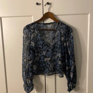 Blommig blus från Sinsay - Säljer en blommig blus från Sinsay i blått och svart med v-ringning. Blusen har långa ärmar och ett luftigt, lätt material som ger en skön känsla. Perfekt för både vardag och speciella tillfällen.