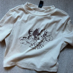Beige ribbad topp med tryck - Säljer en croppad beige ribbad topp med ett tryck av änglar och texten 'ANGEL BABY' på framsidan. Toppen har korta ärmar och en rund halsringning.  