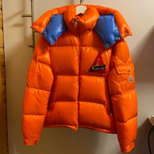 Moncler jacka  - Säljer en cool orange pufferjacka från Moncler. Perfekt för att hålla värmen under kalla dagar. Modellen på jackan heter Wilson och är storlek 2 (M). Extremt bra skick och knappt använd! Ytterst lite smuts på armen som lätt går bort och syns knappt