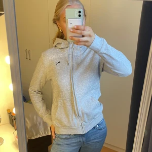 Grå kofta från Puma - Säljer en stilren grå hoodie från Puma med dragkedja och huva. Tröjan har långa ärmar och en liten Puma-logga på bröstet. Den är i storlek S. Kom privat vid intresse!