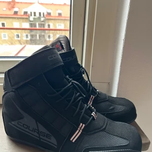 Svarta Course V2 MC boots med snörning - Säljer ett par svarta Course boots med snörning och kardborreband. Skorna har en robust design med en diskret logga på sidan och röd detalj på sulan. Perfekta för en stilren look. Strl 46. Endast använd 1-2 gånger. 