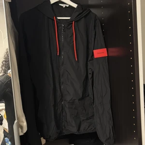 Svart vindjacka från Calvin Klein  - Snygg svart vindjacka från Calvin Klein med huva och röda detaljer. Jackan har dragkedja framtill och en röd logga på ärmen. Perfekt för blåsiga dagar.
