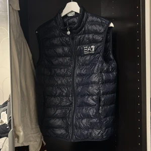 Svart dunväst från EA7 - Snygg svart dunväst från EA7 med dragkedja framtill. Perfekt för kyligare dagar och ger en sportig look. Västen har en quiltad design och är ärmlös, vilket gör den lätt att bära över en tröja eller hoodie.