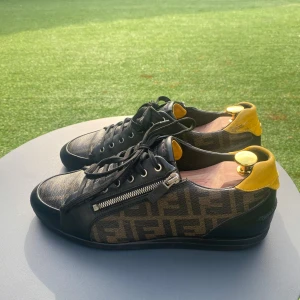 Fendi sneakers storlek 41 - Snygga svarta sneakers med gul häl och mönstrad sida. Skorna har snörning och dragkedja på sidan för extra stil. Perfekta för en trendig look.