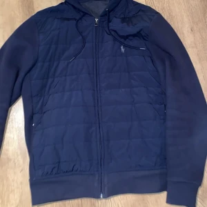 Mörkblå jacka med dragkedja - Snygg mörkblå jacka från Ralph Lauren med dragkedja och huva. Jackan har quiltade detaljer på framsidan och långa ärmar. Perfekt för en stilren look. Strl M