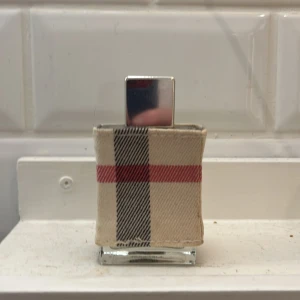 Parfym burberry - Säljer en elegant parfymflaska klädd i ett klassiskt rutigt tygfodral med beige, svart och röd design. Flaskan har en stilren, silverfärgad kork och rymmer 30 ml. Perfekt för den som gillar en tidlös och sofistikerad stil. Nästan full 28ml kvar 