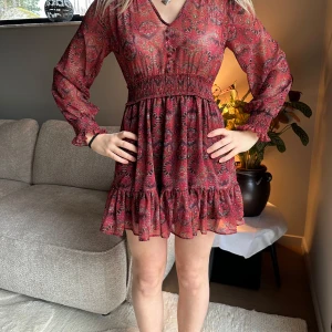 Volang klänning - Charmig röd klänning med ett vackert mönster och volangdetaljer. Den har långa ärmar och en smickrande v-ringning. Perfekt för en bohemisk look.