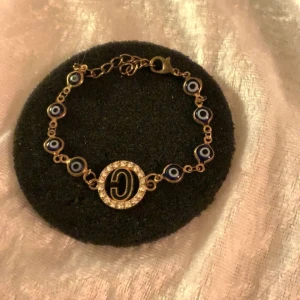 Guldarmband med ögonmotiv och strass - Elegant guldarmband med blå ögonmotiv och en glittrande strassdekoration i mitten. Armbandet har en justerbar kedja och ett stilrent spänne. Perfekt för att ge en touch av glamour till din outfit.