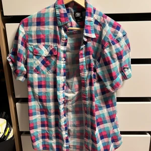 Rutig kortärmad skjorta från FSBN Fishbone - Säljer en färgglad rutig skjorta från Fashion med korta ärmar. Skjortan har ett mönster i blått, rosa och vitt och är perfekt för sommardagar. Den har knappar framtill och två bröstfickor med knappdetaljer.