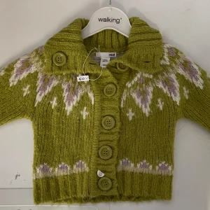 Grön stickad kofta från H&M - Säljer en söt grön stickad kofta storlek 86 från H&M med ett vackert mönster i vitt och lila. Koftan har stora knappar framtill och en mysig krage. Perfekt för kyliga dagar!