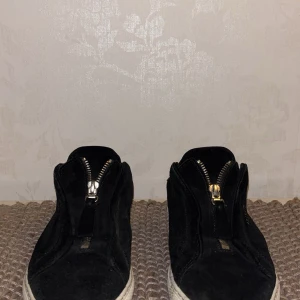 Svarta sneakers från Axel Arigato - Snygga svarta sneakers från Axel Arigato i mocka med dragkedja framtill. Skorna har en vit sula och diskret logga på sidan. Perfekta för en stilren look. Allt og medkommer förutom stickers