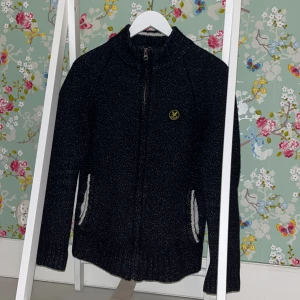 Svart stickad kofta från Lyle & Scott - Snygg svart stickad kofta från Lyle & Scott med dragkedja och ribbade detaljer. Den har en liten logotyp på bröstet och praktiska fickor. Perfekt för kyligare dagar! Sitter perfekt på mig och är ca 174