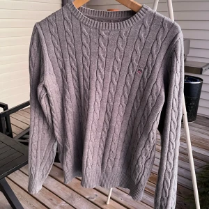 Grå kabelstickad tröja från GANT - Säljer en stilren grå kabelstickad tröja från GANT i premium bomull. Tröjan har en klassisk rund hals och ett diskret broderat märke på bröstet. Perfekt för en avslappnad men ändå elegant look. Väldigt fint skick finns inga defekter. Bara höra av sig vid frågor!😊