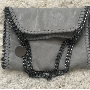 Grå axelväska med kedjedetaljer - Snygg grå axelväska i mocka med kedjedetaljer och leopardmönstrat foder. Väskan har en stilren design med en kedja som axelrem och en dekorativ medaljong. Perfekt för att ge din outfit en edgy touch.