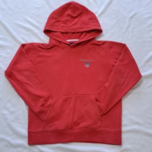 Röd hoodie från GANT - Säljer en snygg röd hoodie från GANT med broderad logga på bröstet. Den har en klassisk känguruficka och ribbade muddar vid ärmslut och nederkant. Perfekt för en avslappnad stil!