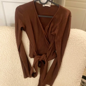 Brun ribbad omlottopp från Zara - Snygg brun ribbad omlottopp från Zara med långa ärmar och knytning i midjan. Perfekt för en stilren look. Passar bra till både jeans och kjol.