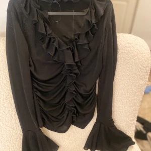 Svart blus med volangdetaljer - Elegant svart blus med volangdetaljer längs framsidan och vid ärmsluten. Blusen har långa ärmar och en vacker drapering som ger en stilfull look. Perfekt för en chic och sofistikerad stil.
