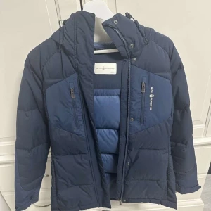 Mörkblå dunjacka från Peak Performance - Säljer en snygg mörkblå dunjacka från Peak Performance. Jackan har en praktisk dragkedja och flera fickor med dragkedjor. Perfekt för kyliga dagar med sin vadderade design och justerbara huva. Storlek xs 