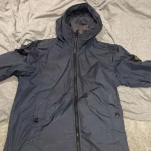 Blå jacka från Stone Island - Säljer en blå jacka från Stone Island med huva och dragkedja. Jackan har långa ärmar och en broderad logotyp på ärmen. Perfekt för en stilren look.