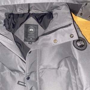 Grå dunjacka från Canada Goose - Säljer en stilren grå dunjacka från Canada Goose med avtagbar huva och dragkedja. Jackan har en broderad logga på ärmen och knappar framtill. Perfekt för kalla vinterdagar.