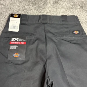 Dickies 874 Byxor - Gråa Dickies workpants i modellen 874 (Original Fit), är både slitstarka och stiliga, perfekt för både arbete och vardagsbruk. De är i helt nyskick, med prislappen kvar, och ordinarie pris är 799 kr. Byxorna erbjuder en bekväm passform och hög kvalitet som kännetecknar Dickies, vilket gör dem till ett pålitligt val. Storlek 30x32.