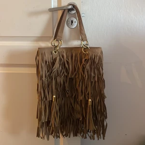 Brun axelväska med fransar - Säljer en snygg brun axelväska med fransar. Väskan har två handtag och är dekorerad med guldfärgade detaljer. Insidan är rymlig med flera fack för enkel organisering. Perfekt för att ge din outfit en bohemisk touch. Äkta Mocca