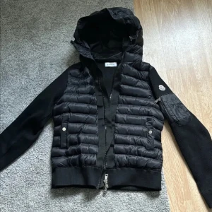 Svart pufferjacka från Moncler - Snygg svart pufferjacka från Moncler med huva och dragkedja. Jackan har långa ärmar och en quiltad design. Perfekt för kyligare dagar med stil.