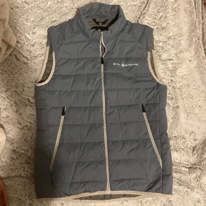 Sail Racing dunväst - Snygg grå dunväst från Sail Racing med dragkedja och två praktiska fickor framtill. Perfekt för kyligare dagar och ger en sportig look. Västen har en stilren design med logotyp på bröstet och nacken.