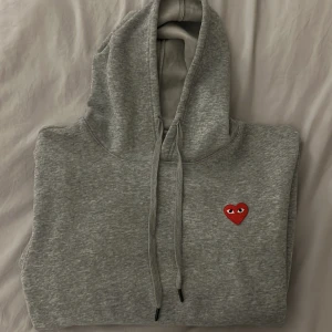 Grå CDG hoodie med rött hjärta  - Säljer en stilren grå cdg hoodie med ett rött hjärta på bröstet. Hoodien har en klassisk design med luva. Sicket är 10/10 för att jag har bara använt den ett få tal gånger! Otroligt snygg nu till våren!🤩men vet inte om den är äkta!