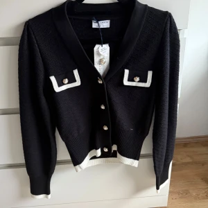 Svart kofta med vita detaljer från Lindex  - Snygg svart kofta från Lindex med vita detaljer och pärlknappar. Koftan har långa ärmar och ribbade muddar. Perfekt för en stilren look.