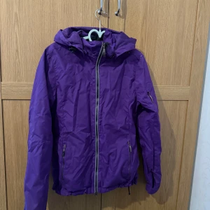 Lila vindjacka med huva - Säljer en snygg lila vindjacka med huva och dragkedja framtill. Jackan har praktiska fickor med dragkedjor och justerbar nederkant. 