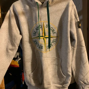Grå hoodie från Stone Island - Säljer en grå hoodie från Stone Island med det ikoniska kompassmärket broderat på bröstet. Hoodien har gröna dragsnören och en praktisk magficka. Barnstorlek M/ motsvarande S i vuxen 