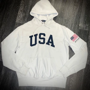 Vit hoodie från Polo Ralph Lauren - Säljer en vit hoodie från Polo Ralph Lauren med 'USA' tryckt i mörkblått på framsidan. Den har en känguruficka och en amerikansk flagga med 'Polo' på ärmen. Perfekt för en avslappnad stil.