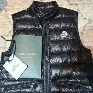 Svart dunväst från Moncler - Snygg svart dunväst från Moncler med dragkedja och hög krage. Västen har ett stilrent quiltat mönster och Monclers logotyp på bröstet. Perfekt för kyligare dagar.