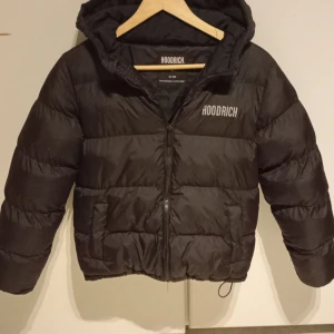 Svart pufferjacka från Hoodrich - Säljer en svart pufferjacka från Hoodrich med huva och dragkedja. Jackan har märkets logga både fram och bak, samt praktiska fickor. Perfekt för kyligare dagar. Jackan har inga fläckar som inte går att ta bort.