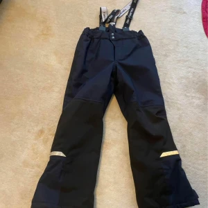 Svarta vinterbyxor - Svarta täckbyxor från Polarn O. Pyret med justerbara hängslen och reflexdetaljer på benen. Perfekta för vinteraktiviteter med slitstarkt material och bekväm passform.