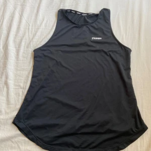 Svart linnetopp från Stronger - Säljer en svart linnetopp från Stronger. Den är ärmlös och perfekt för träning. Materialet är mjukt och stretchigt, vilket gör den bekväm att bära under intensiva pass. Toppen har en enkel och stilren design med 'Stronger' tryckt på bröstet.